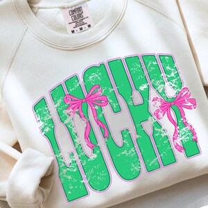 May include: Cream-colored sweatshirt with the word "LUCKY" in green block letters outlined in pink. The word "LUCKY" has a distressed texture and is decorated with pink bows. The sweatshirt has a crew neck and long sleeves.
