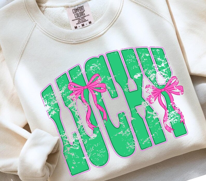 May include: Cream-colored sweatshirt with the word "LUCKY" in green block letters outlined in pink. The word "LUCKY" has a distressed texture and is decorated with pink bows. The sweatshirt has a crew neck and long sleeves.