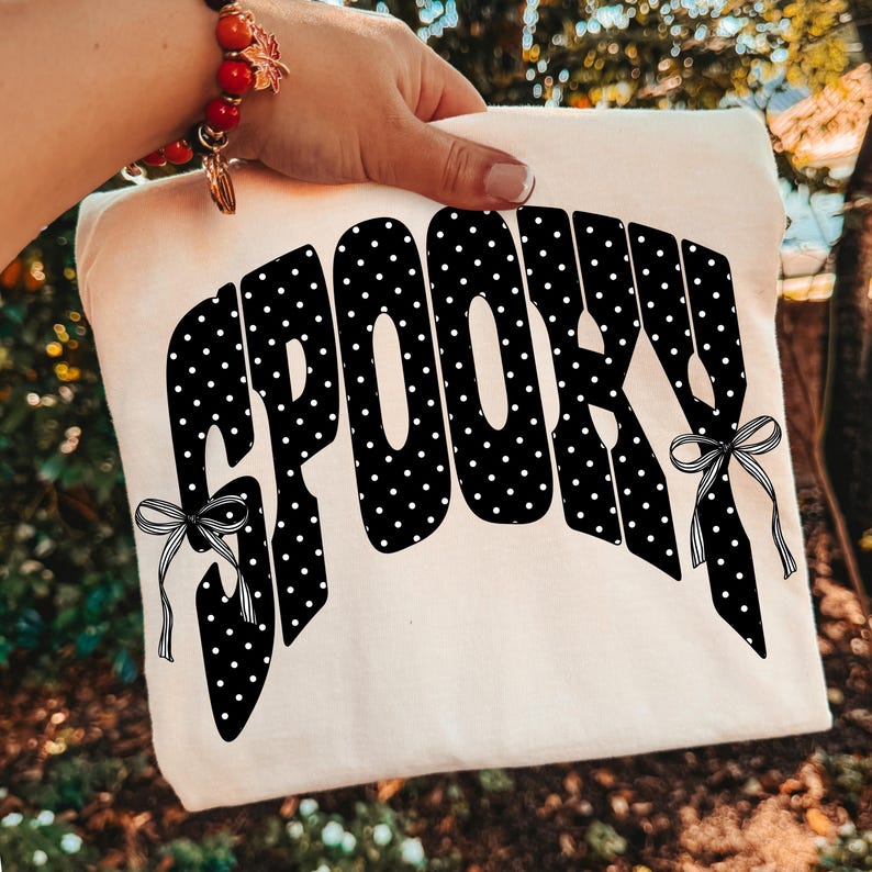 K&ouml;nnte beinhalten: Ein cremefarbenes T-Shirt mit dem Wort SPOOKY in einer gro&szlig;en, geschwungenen schwarzen Schrift mit wei&szlig;en Punkten. Die Buchstaben sind mit zwei schwarz-wei&szlig; gestreiften Schleifen verziert, die sich links und rechts vom Text befinden.