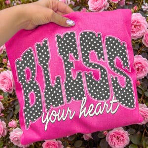 May include: A hot pink sweatshirt with the words "BLESS your heart" in a retro font. The letters are filled with a black and white polka dot pattern. The sweatshirt is held up by a hand, surrounded by pink roses.
