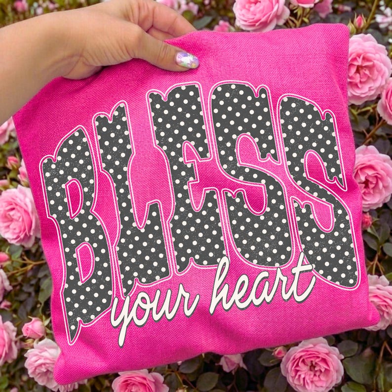 May include: A hot pink sweatshirt with the words "BLESS your heart" in a retro font. The letters are filled with a black and white polka dot pattern. The sweatshirt is held up by a hand, surrounded by pink roses.