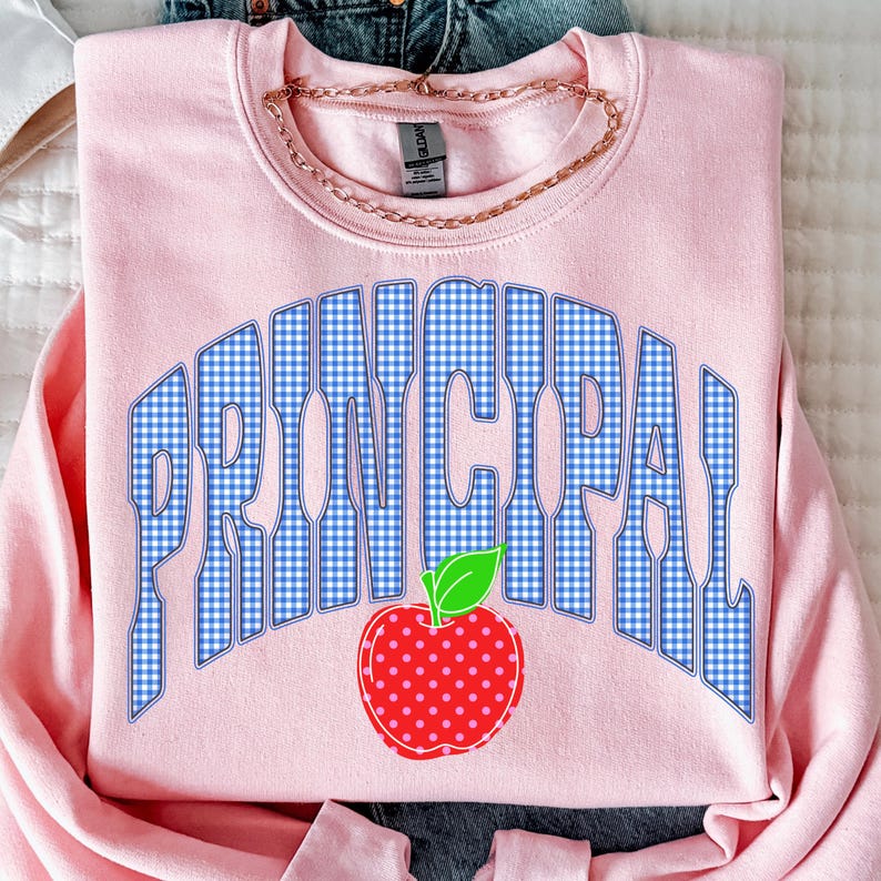 以下が含まれることがあります： ライトピンクのクルーネックトレーナー。青と白のギンガムチェック柄で「PRINCIPAL」という文字がアーチ状に描かれ、その下に白い水玉模様の赤いリンゴのグラフィックが配置されています。襟元はリブ編みで、ブランドタグが見えています。