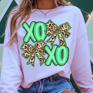 May include: A light pink sweatshirt featuring the letters "XO" in a green bubble font, with leopard print bows. The design is centered on the front of the shirt, creating a playful and stylish look.