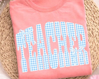 May include: A coral-colored t-shirt with the word "TEACHER" in large, light blue gingham letters. The word has a bow on the "T" and the "R". The shirt is folded on a woven mat with pink flowers.