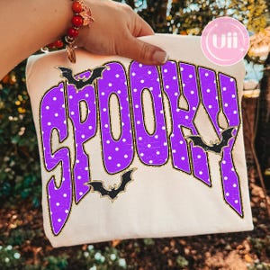 Op de afbeelding: Een crèmekleurig T-shirt met het woord SPOOKY in grote, paarse letters met witte stippen en een verweerde textuur. De tekst is omlijnd met gouden glitters en versierd met drie zwarte vleermuissilhouetten rondom de letters.