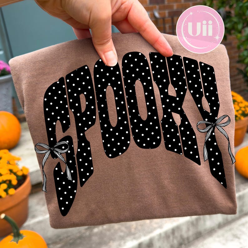K&ouml;nnte beinhalten: Ein braunes T-Shirt mit dem Wort SPOOKY in gro&szlig;en, fetten, schwarzen Buchstaben mit wei&szlig;em Punktmuster. Die Buchstaben sind mit kleinen, schwarz-wei&szlig; gestreiften Schleifen verziert, die an den &auml;u&szlig;eren Enden des Wortes befestigt sind.