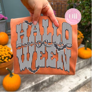 May include: An orange t-shirt featuring the text HALLOWEEN in a bold, black and white vertical striped font. The design includes a central black bow graphic and three black bat silhouettes positioned around the lettering on the front of the shirt.