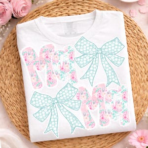 May include: White t-shirt with the word "MOM" in a floral design with pink and blue bows. The letters are filled with a floral pattern of pink and white flowers. The bows are light blue with white polka dots.