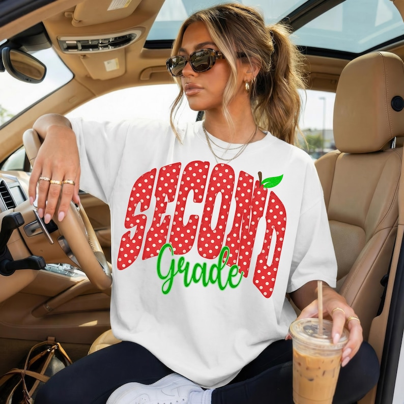 May include: A white short-sleeved t-shirt featuring a large red graphic with white polka dots that spells out the text SECOND in a bold font, with the word Grade written below in green cursive script and a small green apple icon attached.