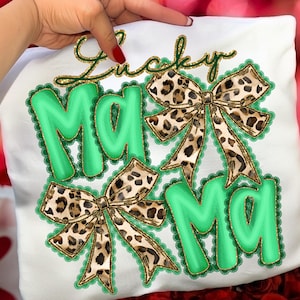 May include: White t-shirt with the words "Lucky Mama" in green and gold script. The word "Mama" is in large green letters with a scalloped border, and leopard print bows are featured.