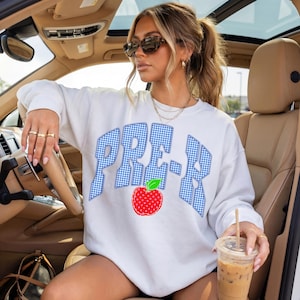 May include: White sweatshirt with the word "PRE-K" in blue gingham letters and a red apple graphic. The sweatshirt is a casual style, perfect for a teacher or student. The image is taken inside a car.