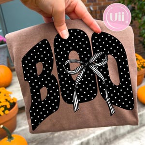 Puede incluir: Una camiseta marrón con la palabra BOO en letras negras grandes con un patrón de lunares blancos. Un lazo a rayas blancas y negras está atado alrededor de la letra O central. El diseño está impreso en la parte delantera con estilo gráfico.