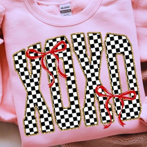 Puede incluir: Una sudadera rosa claro con las letras "XOXO" en un patr&oacute;n de cuadros blancos y negros. Las letras est&aacute;n delineadas con purpurina dorada y adornadas con lazos rojos. La sudadera tiene cuello redondo y mangas largas.