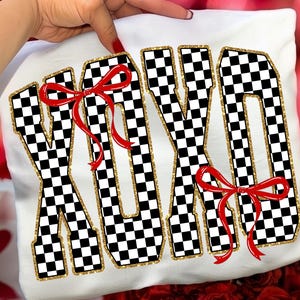 Puede incluir: Camiseta blanca con las letras XOXO en un patr&oacute;n de cuadros blanco y negro, delineadas con purpurina dorada. Lazos rojos est&aacute;n unidos a las letras. La camiseta es sostenida por una mano.