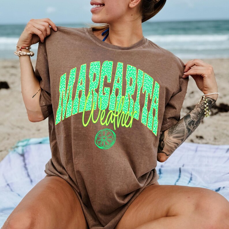 May include: Brown t-shirt with the words "MARGARITA Weather" in bright green, with a lime graphic. The shirt is worn on a beach, suggesting a summer or vacation theme. The text is in a bold, playful font.