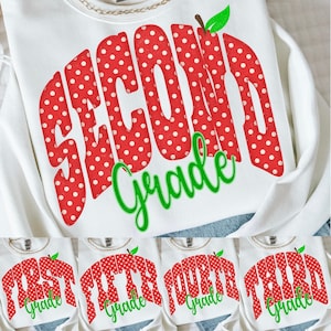May include: A white sweatshirt featuring a large red graphic with white polka dots that spells out grade levels like Second Grade, First Grade, Third Grade, Fourth Grade, or Fifth Grade. A small green leaf sits atop the red text.