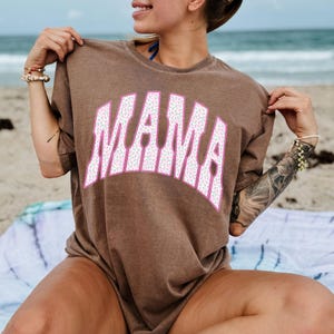 Peut inclure: Un t-shirt marron avec le mot "MAMA" en grandes lettres blanches &agrave; imprim&eacute; l&eacute;opard, bord&eacute;es de rose. Le t-shirt est port&eacute; par une personne sur une plage, avec l'oc&eacute;an en arri&egrave;re-plan. Le t-shirt a des manches courtes et une coupe ample.
