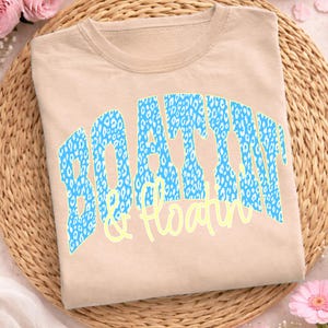 May include: A beige t-shirt featuring the text "BOATTIN & Floatin". The word "BOATTIN" is in a blue leopard print, and "& Floatin" is in a light yellow script. The shirt is folded on a woven mat.