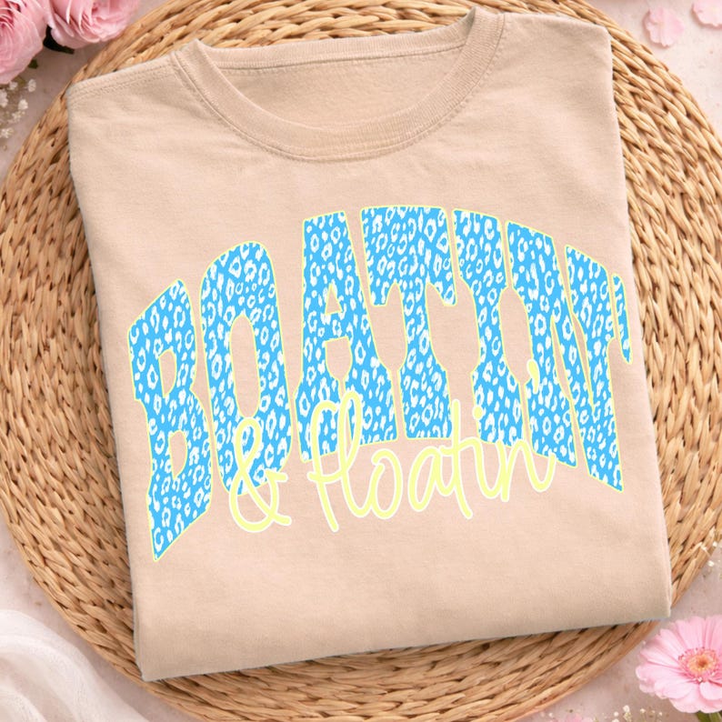 May include: A beige t-shirt featuring the text "BOATTIN & Floatin". The word "BOATTIN" is in a blue leopard print, and "& Floatin" is in a light yellow script. The shirt is folded on a woven mat.