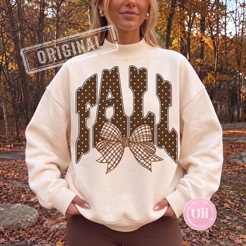 May include: A cream-colored mock neck sweatshirt featuring the word FALL in large, brown, white-polka-dotted letters. A brown and white gingham patterned bow is centered below the text. The sweatshirt has long sleeves and ribbed cuffs.