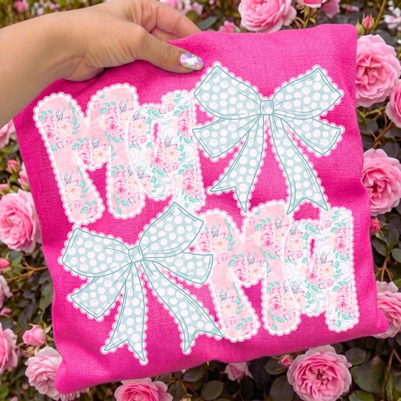 May include: A pink pillow with the word "MOM" in a floral pattern, accented with light blue polka dot bows. The pillow is held up by a hand, with a background of pink roses.
