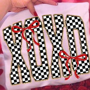 Puede incluir: Camiseta blanca con las letras XOXO en un patr&oacute;n de cuadros blanco y negro, contorneadas con purpurina dorada. Lazos rojos adornan las letras. La camiseta est&aacute; sujeta por una persona.