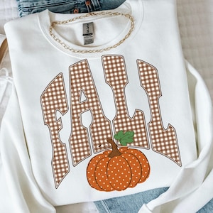 Peut inclure: Un sweat-shirt blanc &agrave; col rond arborant le mot FALL en grandes lettres &agrave; motif vichy marron et blanc. Sous le texte se trouve une citrouille orange &agrave; pois blancs avec une feuille verte. Une cha&icirc;ne dor&eacute;e repose sur le col.