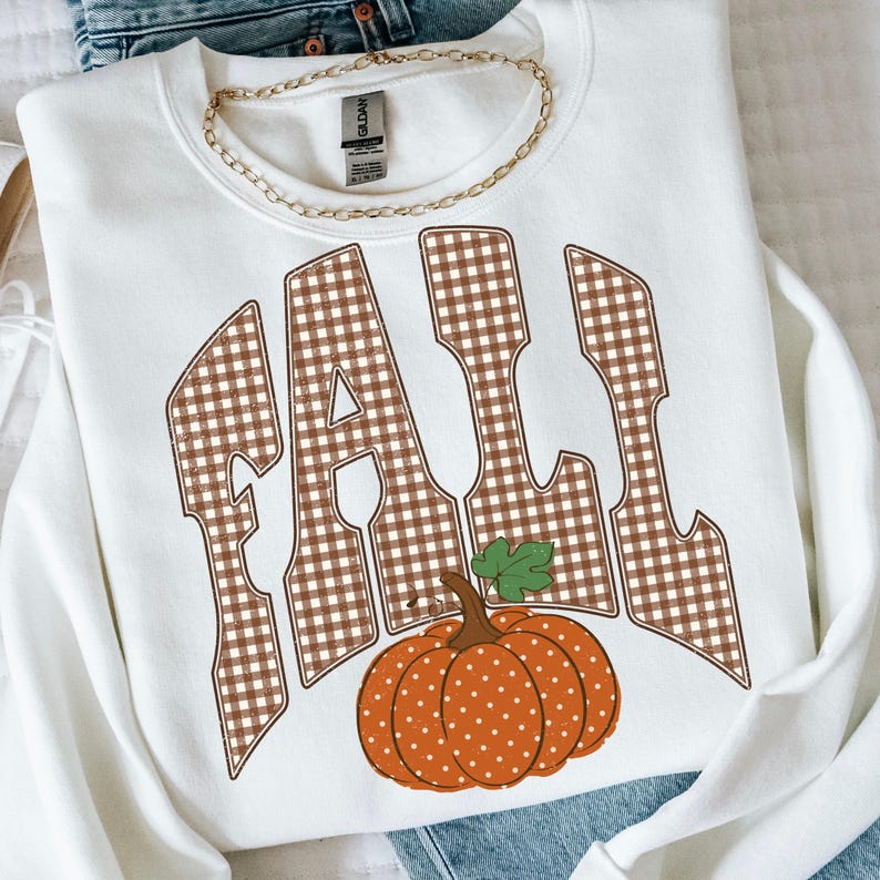Peut inclure: Un sweat-shirt blanc &agrave; col rond arborant le mot FALL en grandes lettres &agrave; motif vichy marron et blanc. Sous le texte se trouve une citrouille orange &agrave; pois blancs avec une feuille verte. Une cha&icirc;ne dor&eacute;e repose sur le col.
