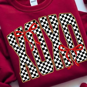 Puede incluir: Una sudadera granate con las letras "XOXO" en un llamativo dise&ntilde;o de cuadros blancos y negros, con ribete de purpurina dorada. Cada letra est&aacute; adornada con un lazo rojo. Una prenda c&oacute;moda y festiva.
