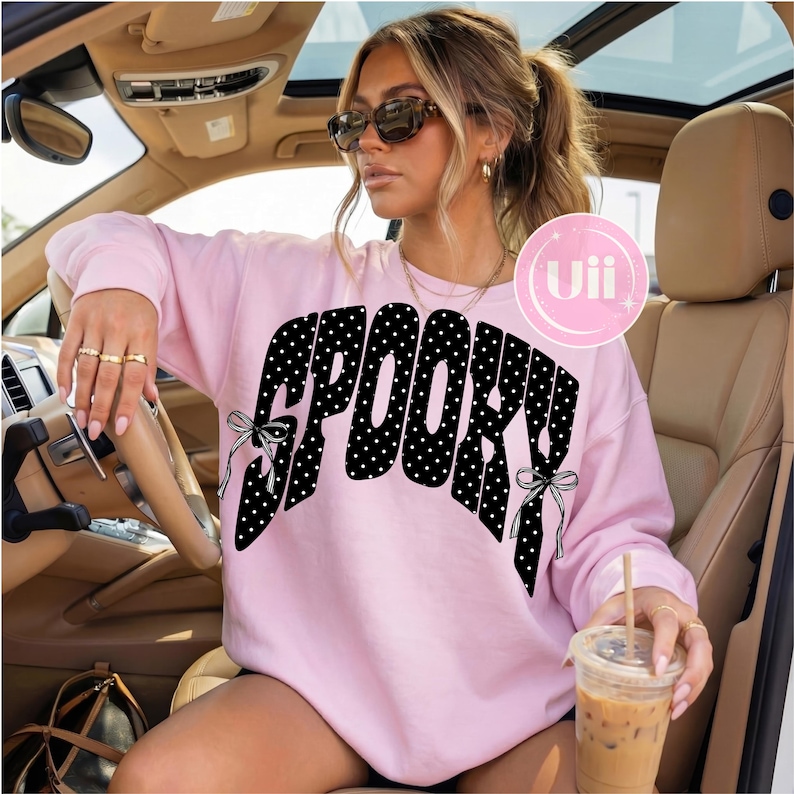 K&ouml;nnte beinhalten: Ein hellrosa Langarm-Sweatshirt mit dem Wort SPOOKY in einer gro&szlig;en schwarzen Schriftart mit wei&szlig;en Punkten. Die Buchstaben sind an den Seiten mit kleinen schwarz-wei&szlig;en Schleifen verziert. Ein kreisf&ouml;rmiges rosa Logo mit dem Text Uii ist sichtbar.