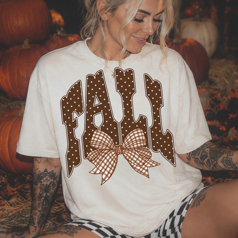 May include: A cream-colored short-sleeve t-shirt featuring the word FALL in large, brown, white-polka-dotted letters. A brown and white gingham patterned bow is centered at the bottom of the text design, layered over the letters.