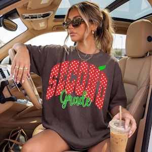 May include: A dark brown short-sleeve t-shirt featuring a large red apple graphic with white polka dots. The text "SECOND" is printed in white on the apple, and the word "Grade" is written below in a bright green cursive script font.