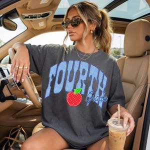 May include: Dark gray t-shirt with the words "FOURTH Grade" in a blue gingham pattern. A red apple graphic with a green leaf is centered below the word "FOURTH". The shirt is worn by a person in a car.