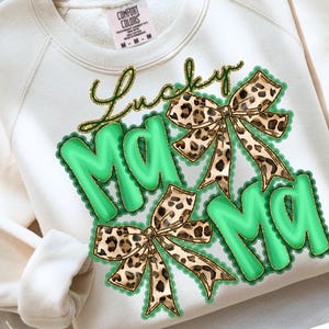 May include: Cream-colored sweatshirt with the words "Lucky Mama" in gold script. The word "Mama" is in large, green, bubble letters with a scalloped border. Two leopard print bows are also featured.