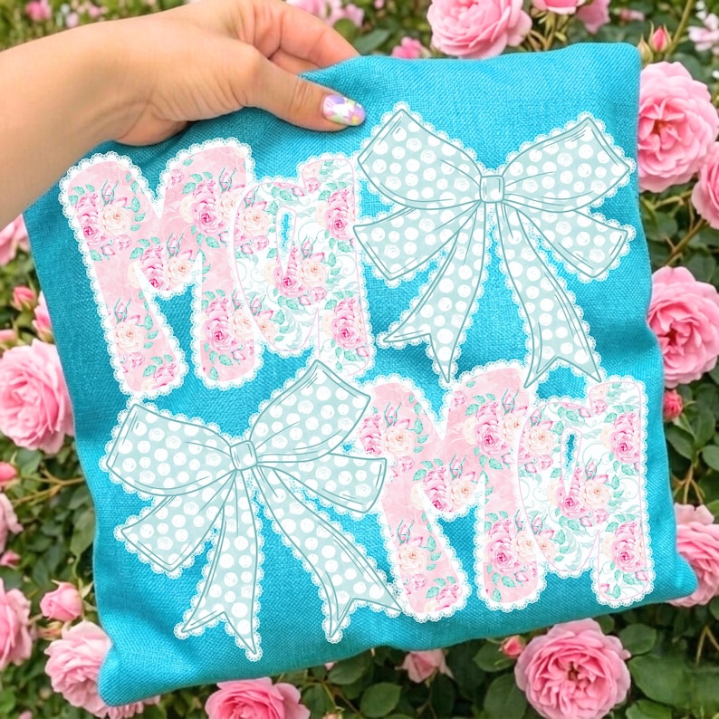 May include: A turquoise pillow with the word "MOM" in pink floral letters and two light blue polka dot bows. The pillow is held in a person's hand, surrounded by pink roses.