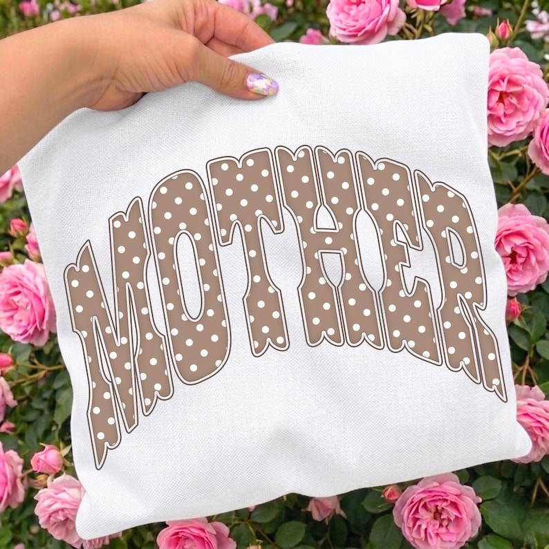 May include: White square pillow with the word "MOTHER" in brown, polka-dotted letters. The pillow is held in front of a background of pink roses.