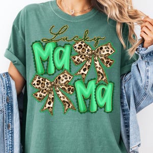 May include: Green t-shirt with the text "Lucky Mama" in gold script and green bubble letters. The design includes two leopard print bows. The shirt is a comfortable, casual style.