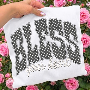 May include: A white pillow with the words "BLESS your heart" in a black polka dot design. The pillow is held by a hand and surrounded by pink roses.