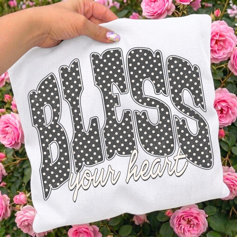 May include: A white pillow with the words "BLESS your heart" in a black polka dot design. The pillow is held by a hand and surrounded by pink roses.