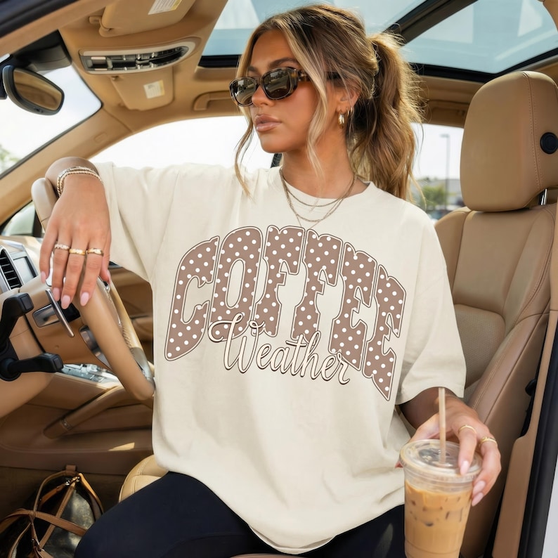 May include: Cream-colored t-shirt with the words "COFFEE Weather" in a brown polka-dot design. The shirt is worn by a person in a car, holding a coffee drink. The shirt is a casual, everyday wear item.