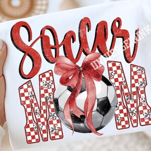 May include: White t-shirt with the words "Soccer Mom" in red script and checkered letters. A soccer ball with a red bow is in the center. The design is a festive, sporty theme.