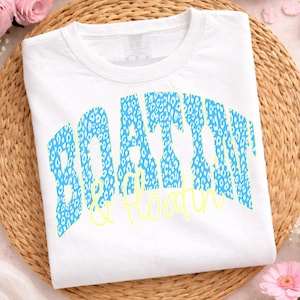 May include: White t-shirt featuring the words "BOATING & FLOATIN" in a blue leopard print design. The words are arched across the front of the shirt, with the word "&" in a yellow script font.