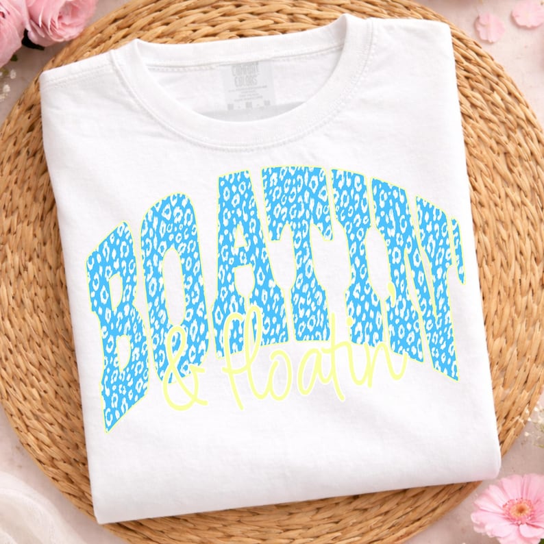 May include: White t-shirt featuring the words "BOATING & FLOATIN" in a blue leopard print design. The words are arched across the front of the shirt, with the word "&" in a yellow script font.
