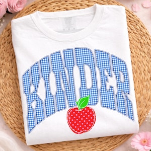 May include: White t-shirt with the word "KINDER" in blue gingham letters and a red apple with pink polka dots. The shirt is a crew neck and is displayed on a woven mat.
