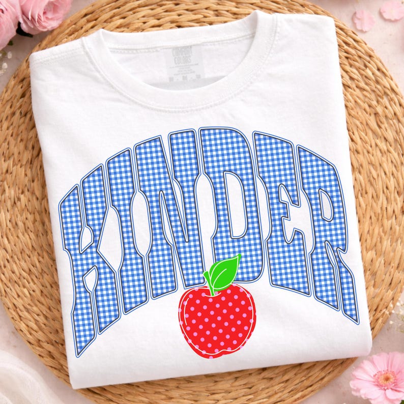 May include: White t-shirt with the word "KINDER" in blue gingham letters and a red apple with pink polka dots. The shirt is a crew neck and is displayed on a woven mat.