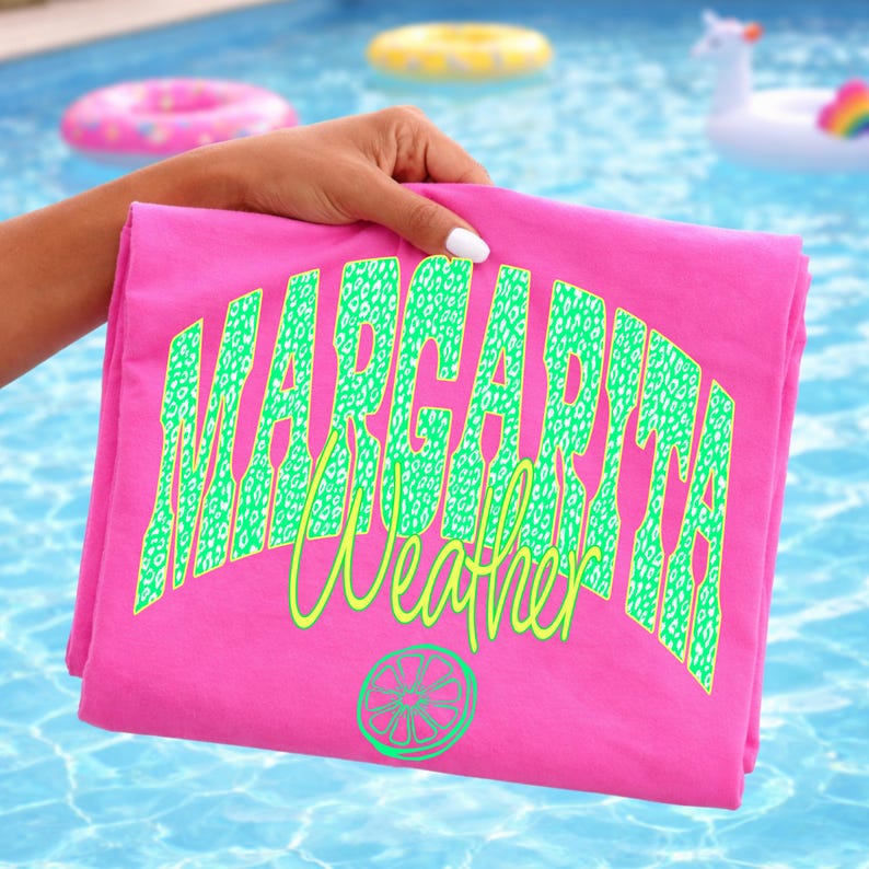 May include: A folded pink t-shirt with the words "Margarita Weather" in green leopard print lettering. A lime slice graphic is below the text. The background shows a swimming pool with inflatable pool toys.