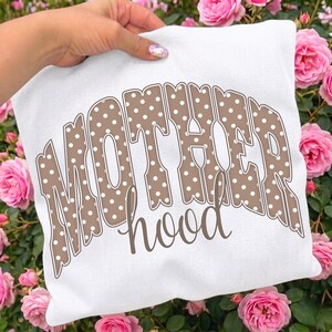 May include: A white square pillow with the word "MOTHERhood" printed on it. The word "MOTHER" is in large, brown, polka-dotted letters, and "hood" is in a smaller, cursive brown font. The pillow is held in front of pink roses.
