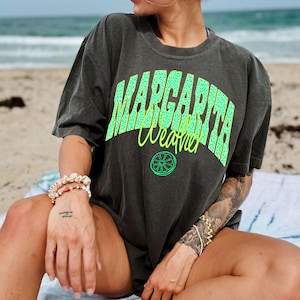 May include: Dark gray t-shirt with the words "MARGARITA Weather" in bright green and yellow. The shirt features a lime graphic and has a relaxed fit. The image is taken on a beach.