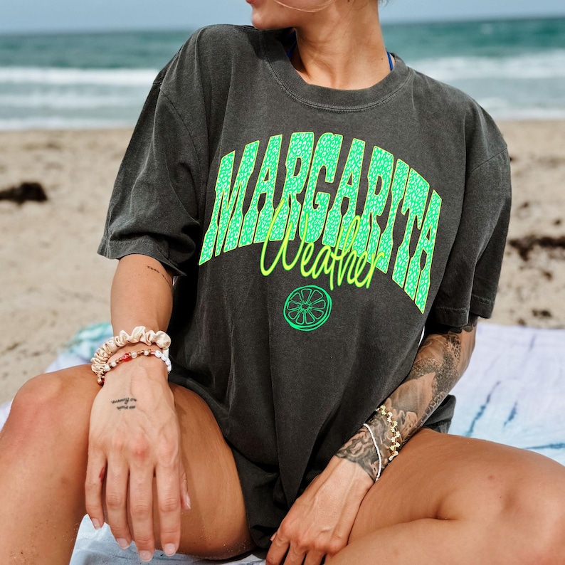May include: Dark gray t-shirt with the words "MARGARITA Weather" in bright green and yellow. The shirt features a lime graphic and has a relaxed fit. The image is taken on a beach.
