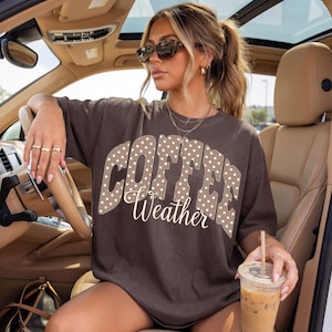 May include: A brown t-shirt with the words "COFFEE Weather" in a white and polka dot design. The person in the image is wearing sunglasses and holding a coffee drink.
